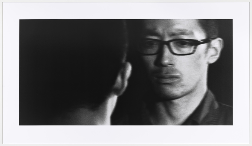 Black and white photo of a man with glasses looking at his reflection with a serious expression.