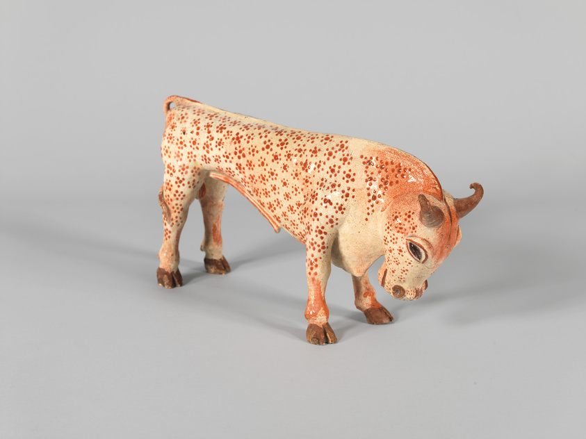 A figurine of a cow with a predominantly white body and numerous small brown spots stands against a plain gray background. The cow has detailed features, including horns, ears, and eyes, and appears to be in a grazing position. The figurine exhibits a rustic texture and a slightly worn appearance, suggesting it may be a vintage or handcrafted item.