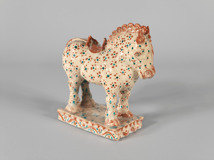 A ceramic figurine of a horse with a speckled pattern of blue and brown dots on a beige background, standing on a rectangular base with similar color splashes. The horse features detailed mane and facial features, with a prominent ridge of mane along its neck highlighted in brown.
