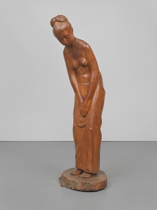 A wooden sculpture of a standing woman with a contemplative expression, bending slightly forward with her hands resting on her thigh. The figure has a detailed hairstyle and is positioned on a circular wooden base against a plain white background.
