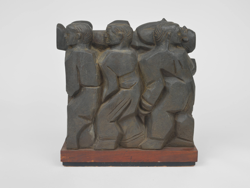 A dark stone sculpture depicting four stylized human figures connected side-by-side, with simplified facial features and geometric body shapes, mounted on a wooden base against a white background.