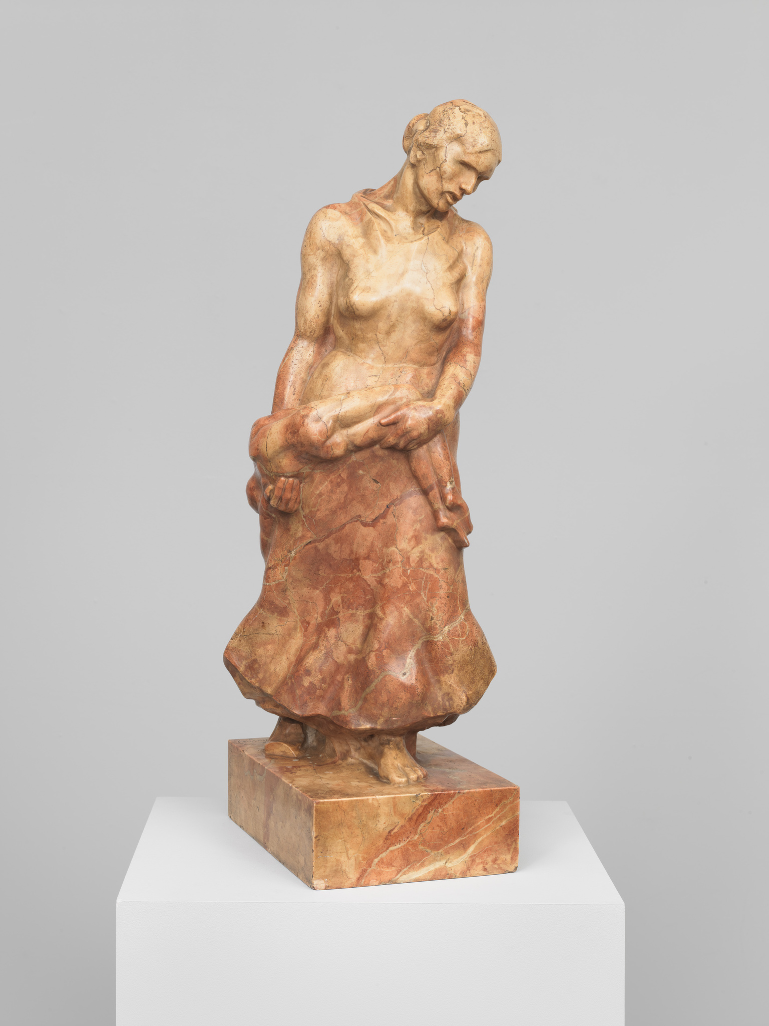 A sculpture of a pensive figure with a draped garment, carved from marble with warm hues, displayed on a white pedestal against a neutral background.