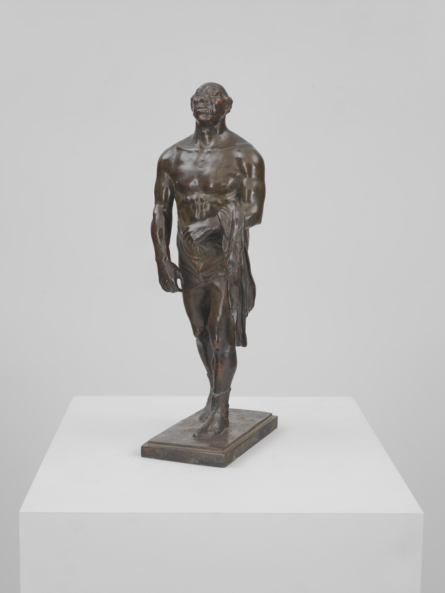 Bronze statue of a standing male figure with a draped cloth, displayed on a white pedestal against a plain background.
