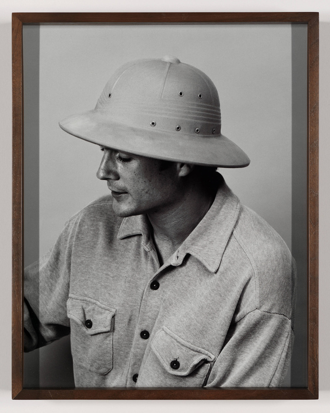 A black and white photograph of a person wearing a wide-brimmed pith helmet and a buttoned-up shirt, looking downward with a contemplative expression. The photo is framed with a simple wooden frame.
