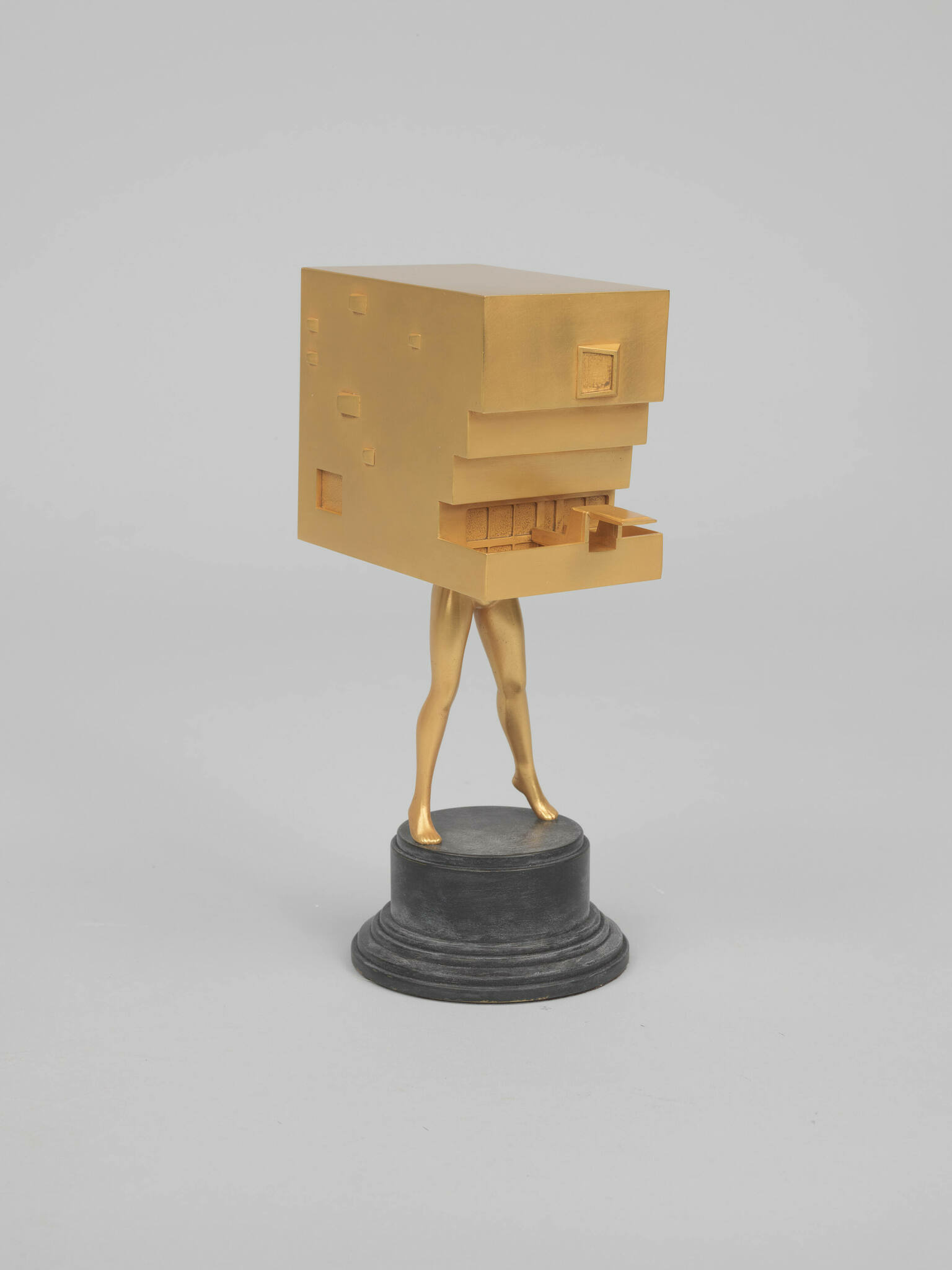 A sculpture featuring golden human-like legs supporting a gold-colored block with architectural details, standing on a round black pedestal against a white background.