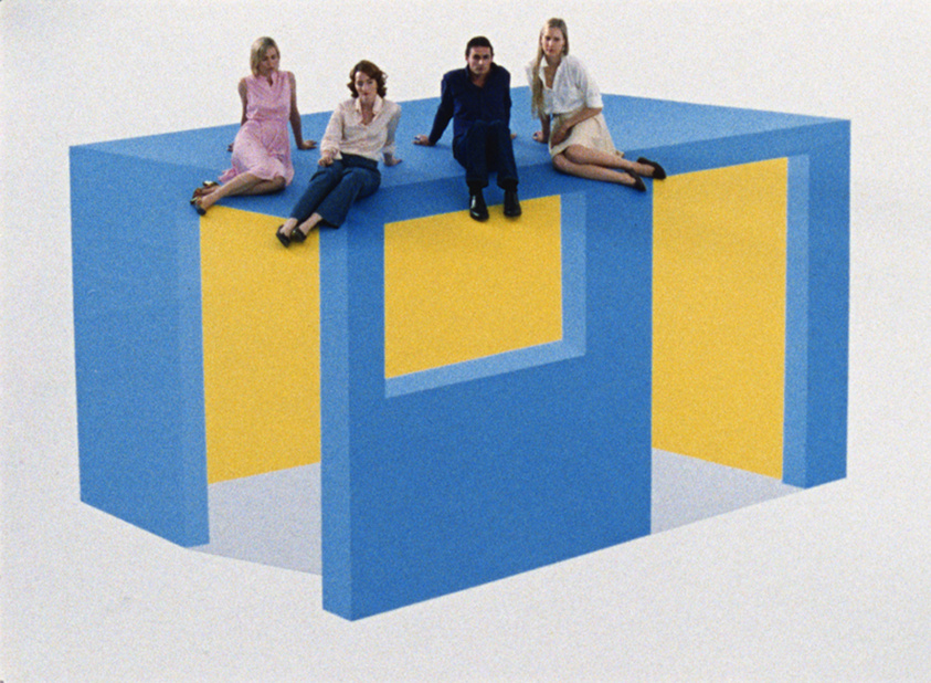 Four people sitting on a large optical illusion artwork that looks like a floating blue and yellow structure.
