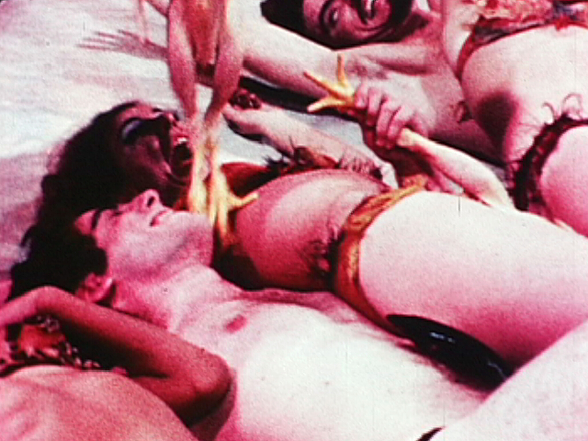 A vintage, grainy photo depicting a scene with three people in what appears to be a playful or theatrical pose. The central figure is lying on their back with their mouth open, while the other two, wearing costumes with feathered headdresses, are leaning over, playfully interacting with the central figure. The image has a reddish-pink tint, adding to its retro aesthetic.