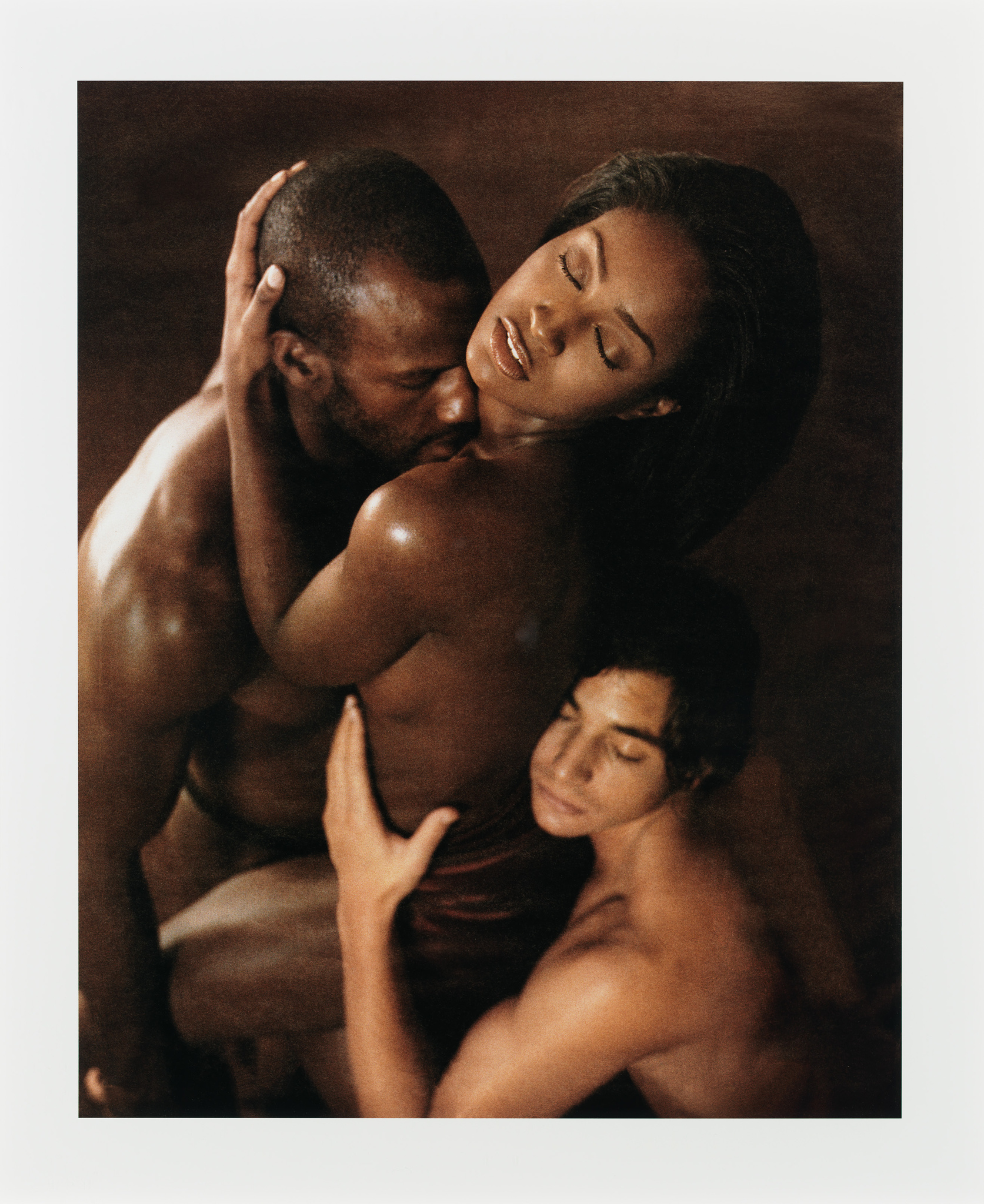 A tender and artistic photograph capturing an intimate moment between three individuals. The image shows two men and a woman in a close embrace, with the woman's head resting gently on one man's shoulder while the other man supports her from beneath. All three subjects are shirtless, highlighting the contours and muscles of their bodies, and they appear to be in a serene state, with their eyes closed and expressions peaceful. The warm, muted tones of the image convey a sense of calm and closeness.