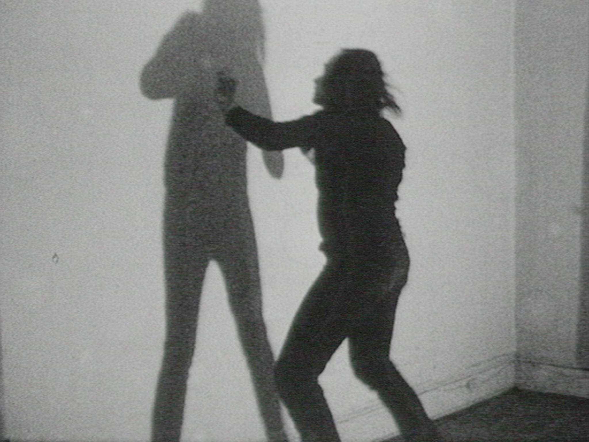 A grainy black and white photo capturing the silhouette of a person against a wall, with their shadow prominently displayed. The person appears to be in motion, possibly dancing or gesturing, with their hair dynamic, suggesting movement.
