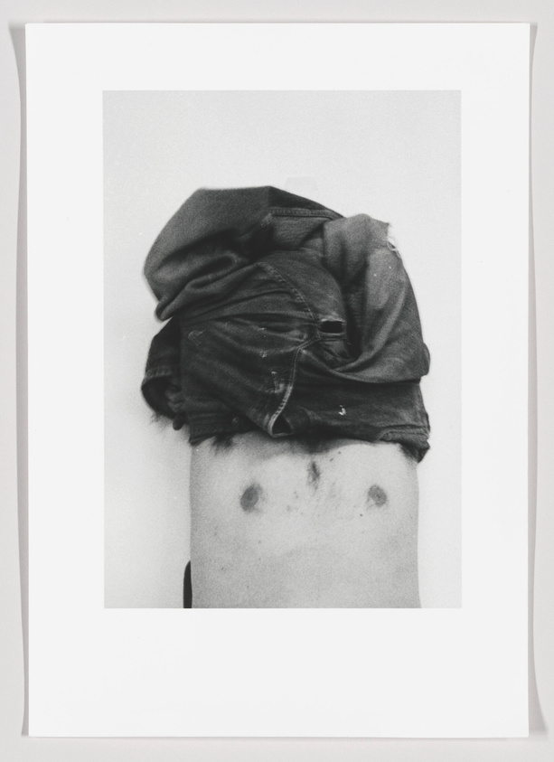 A black and white photograph of a person's torso with a dark shirt lifted up to reveal the stomach area, which has two dark circular marks resembling eyes and a faint line that could suggest a mouth, giving the appearance of a face. The image is framed with a white border.