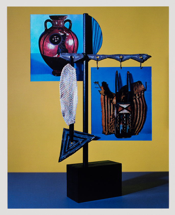 A contemporary art piece featuring a vertical assemblage of various objects and images against a yellow and blue background. The sculpture includes a black stand with a horizontal bar from which a white mesh fabric hangs, and a blue and black triangular form is suspended below. Two printed images are attached to the structure: one of a red vase with a design, and the other of a blue background with a brown object resembling a corset. The piece has a graphic and abstract quality, with a play on textures and shapes.