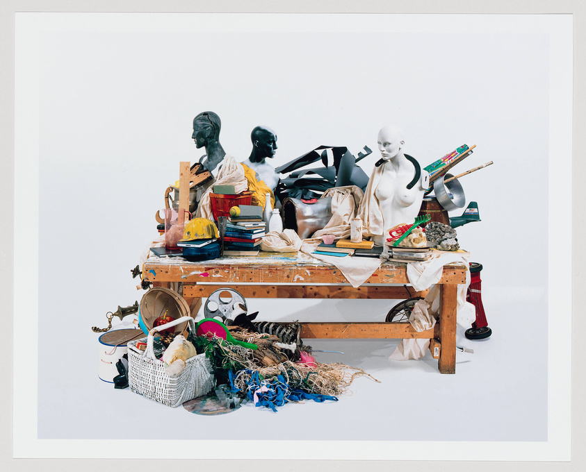 A cluttered workbench filled with an eclectic mix of objects including mannequin heads, books, tools, a hard hat, and various pieces of debris, all against a white background.