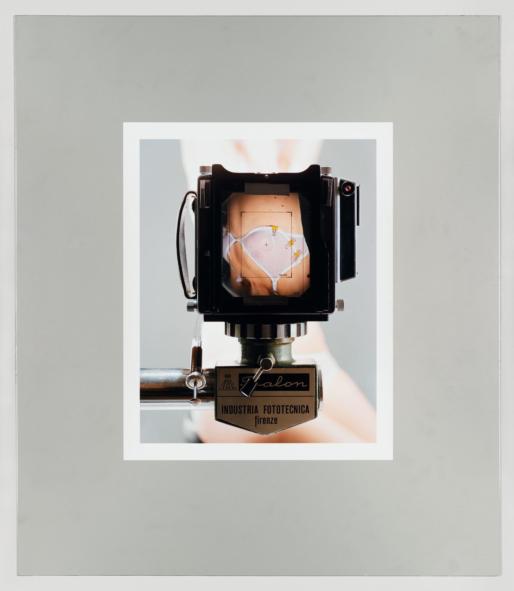 A vintage camera on a tripod is centered in the image, with a close-up view through its viewfinder displaying an upside-down photograph of a person's midsection wearing a shirt with a floral design. The camera is labeled "Falcon" from "Industria Fototecnica Firenze." The background is a neutral gray with a white border framing the camera.