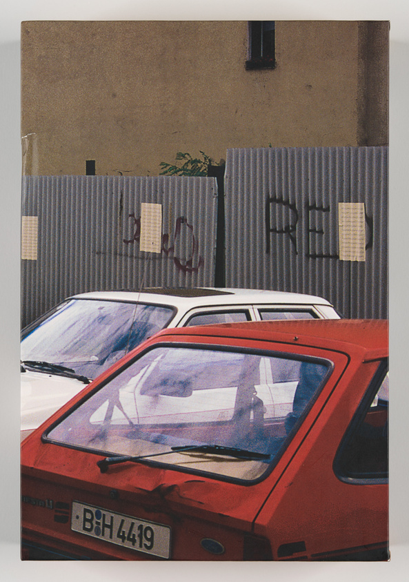 A photograph of a red hatchback car with a European license plate parked in front of a corrugated metal fence with graffiti and taped papers. Behind the fence, a beige building with a small window is visible. Two other cars with blue tones are parked next to the red car, partially visible in the frame.
