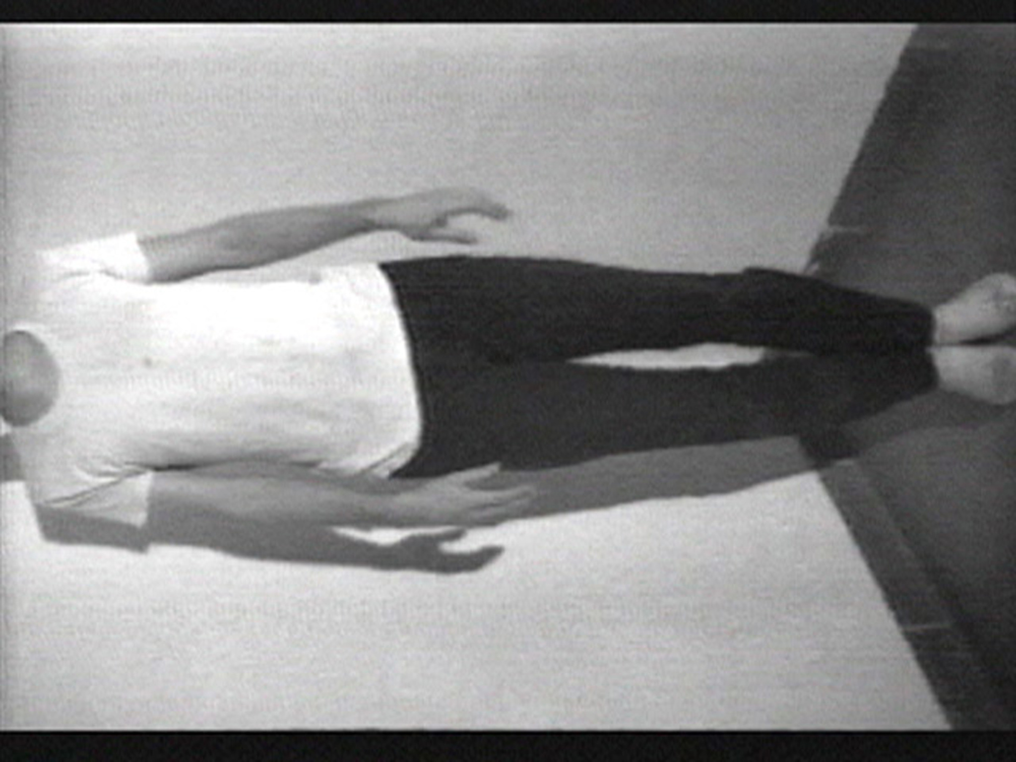 A grainy black and white image of a person lying on their back with arms by their sides on a surface that appears to be sloped or angled, wearing a white t-shirt and dark pants.