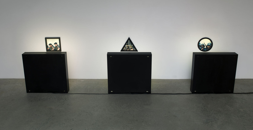 Three black pedestals display framed images: a square, a triangle, and a circle, each illuminated.