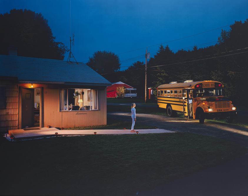 A child in pajamas stands outside a house at night, facing a parked school bus.