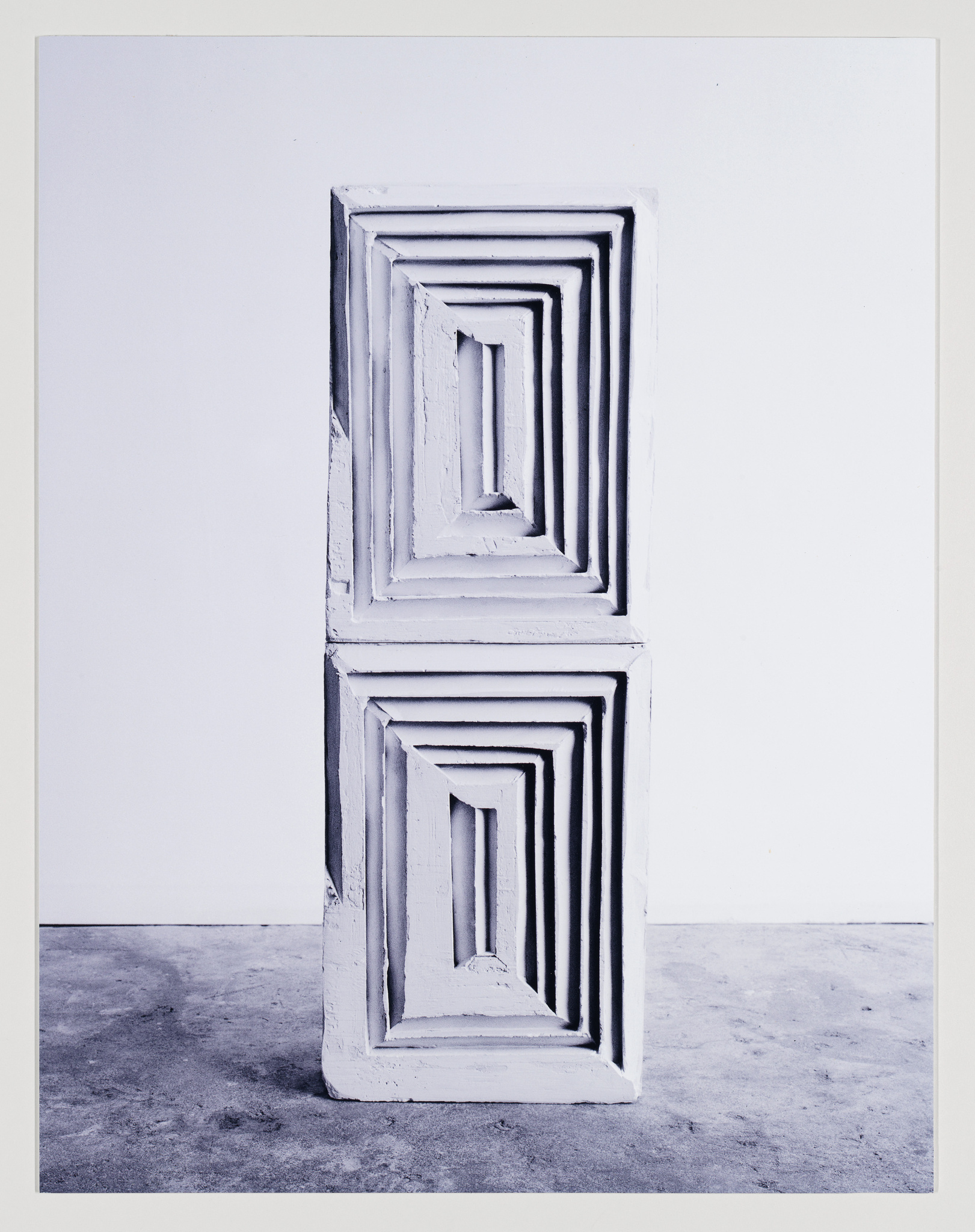 Two white, rectangular frames with a decreasing size pattern stacked vertically on top of each other against a white wall on a concrete floor, creating a visual effect of a tunnel or corridor receding into space.