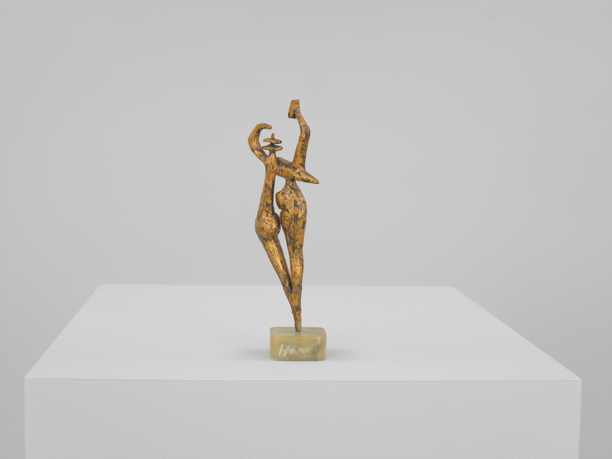 A bronze sculpture of an abstract, elongated figure in a dynamic pose displayed on a white pedestal against a light grey background. The figure appears to be dancing or in motion, with one arm stretched upwards and the other bent at the elbow. The surface of the sculpture has a textured, golden patina.