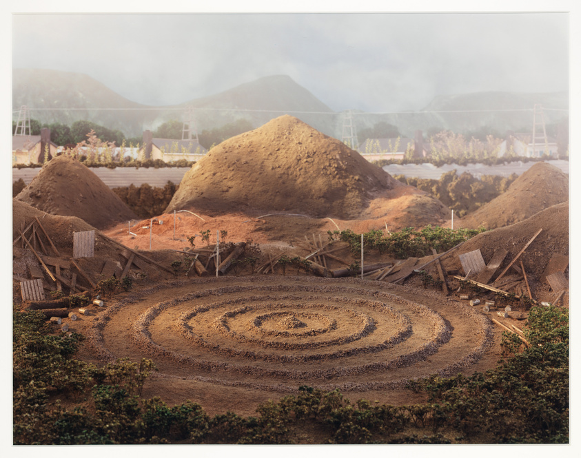 A construction site with several large mounds of dirt and a circular pattern etched into the ground in the foreground. In the background, there is a view of a bridge and hills under a hazy sky. The site appears inactive with no visible workers or heavy machinery.