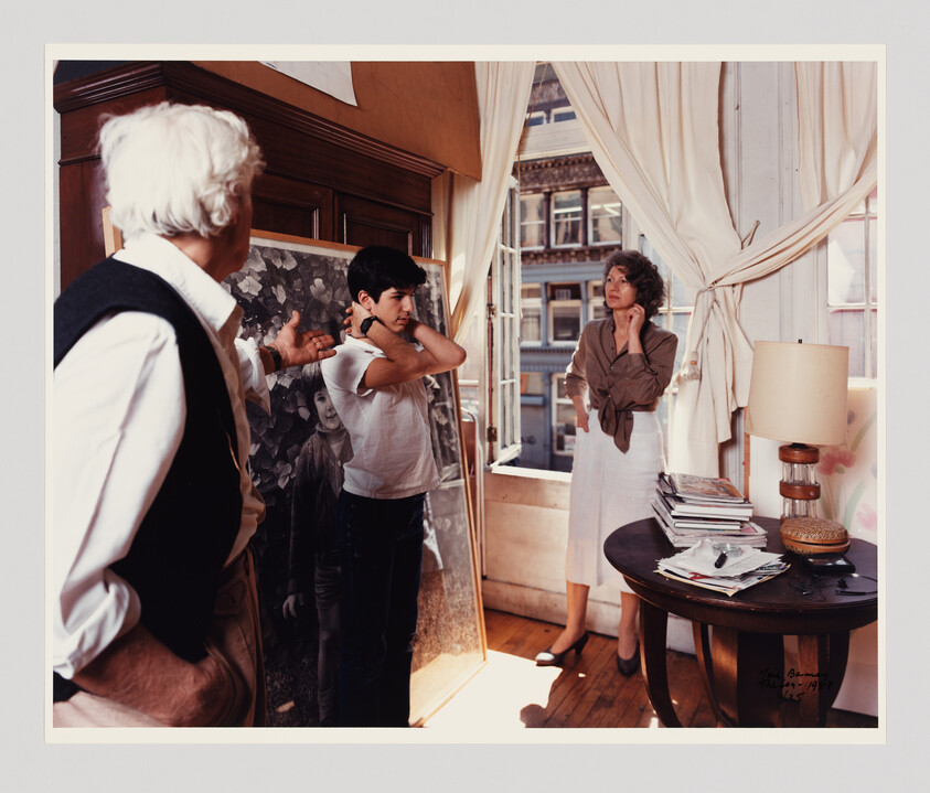 Three people stand in a sunlit room, talking near a window and a table stacked with papers and magazines.
