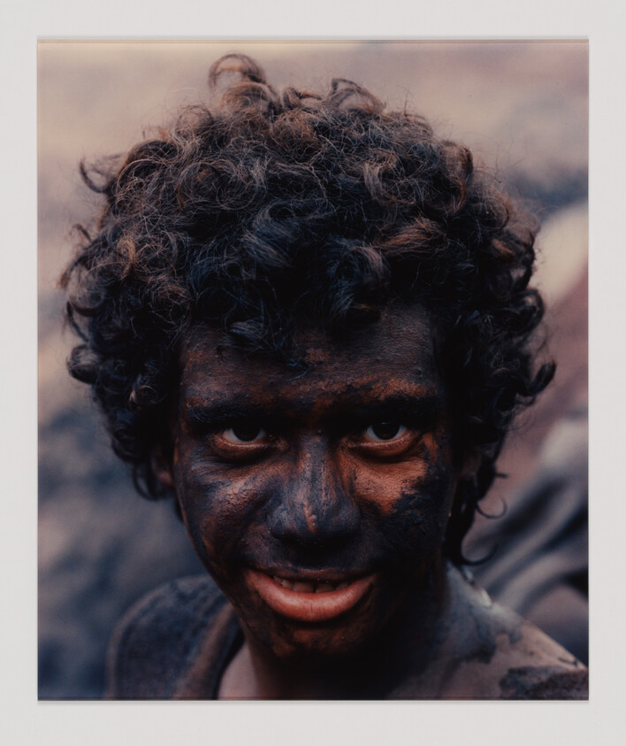 A person with curly hair and a face covered in dark dirt looks directly at the camera, slightly smiling.