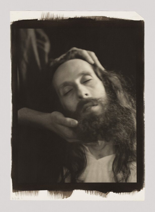 A person with long hair and a beard has their eyes closed while another person gently holds their head.