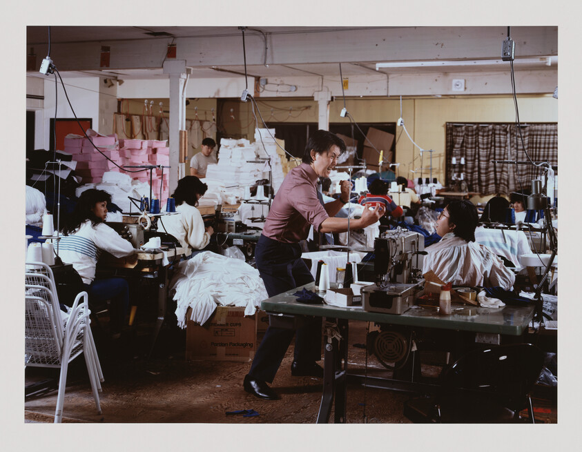 Several people work at sewing machines in a busy garment factory, with one person standing and gesturing to another.