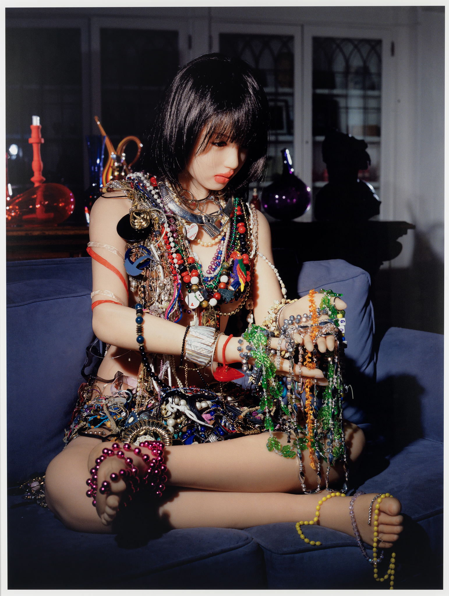 A person with a bob haircut is sitting cross-legged on a blue sofa, covered in an abundance of colorful jewelry, including necklaces, bracelets, and beads. The background features a dimly lit room with glass objects on a table.