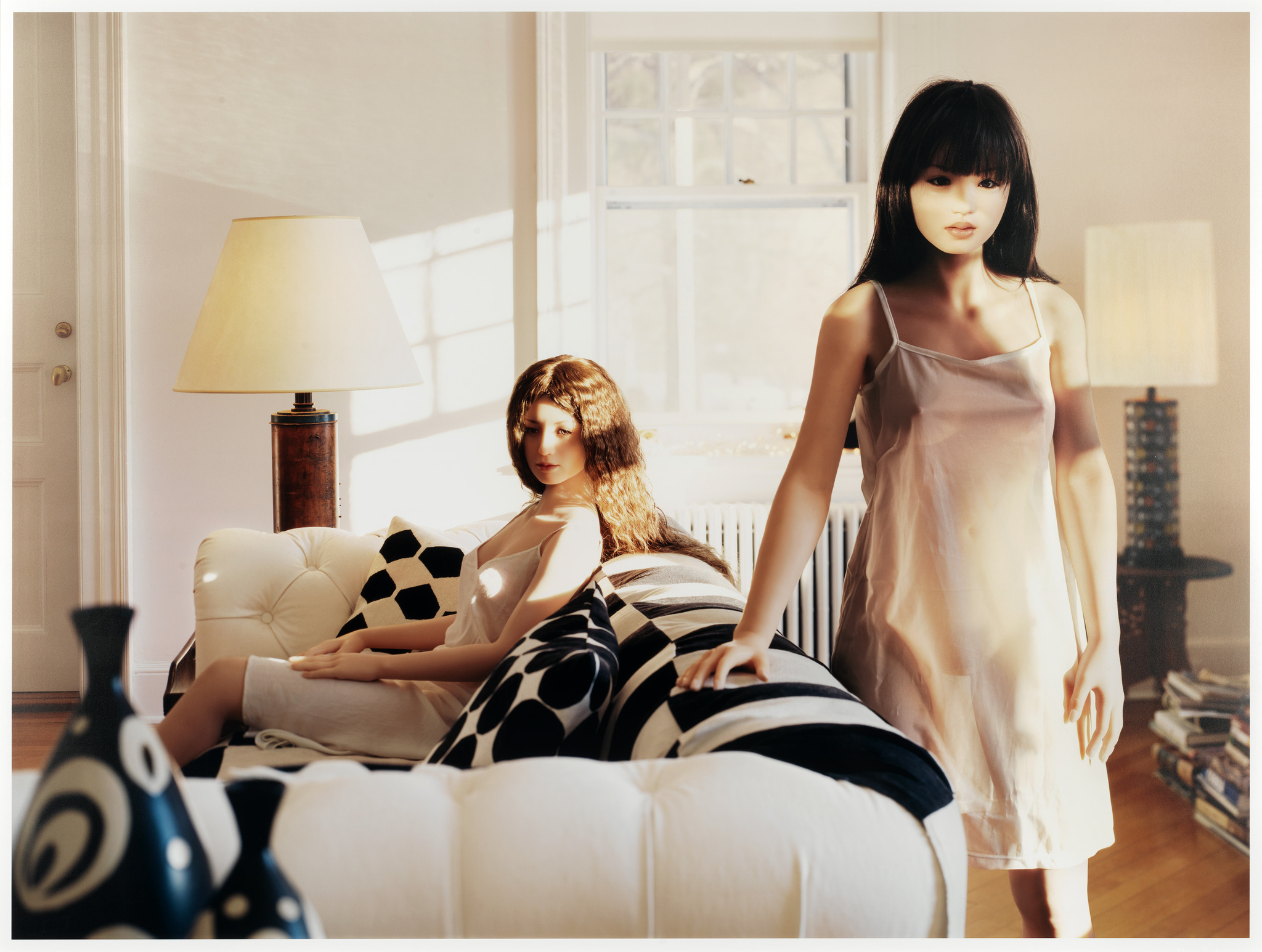 Two mannequins in a sunlit room, one seated on a sofa and the other standing, with a cozy interior setting.