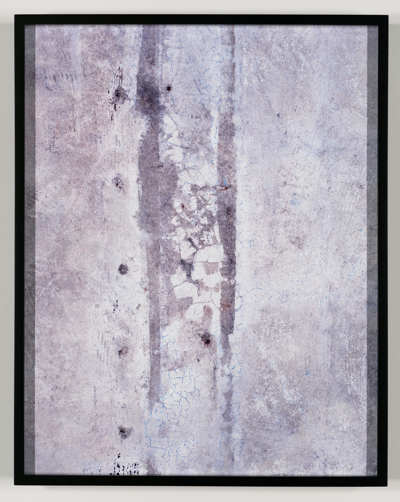 A framed abstract artwork featuring a textured surface with a palette of muted blues, whites, and grays. The composition includes vertical lines and patches of crackled paint, suggesting a weathered wall or surface. The piece is mounted on a wall and encased in a simple black frame.