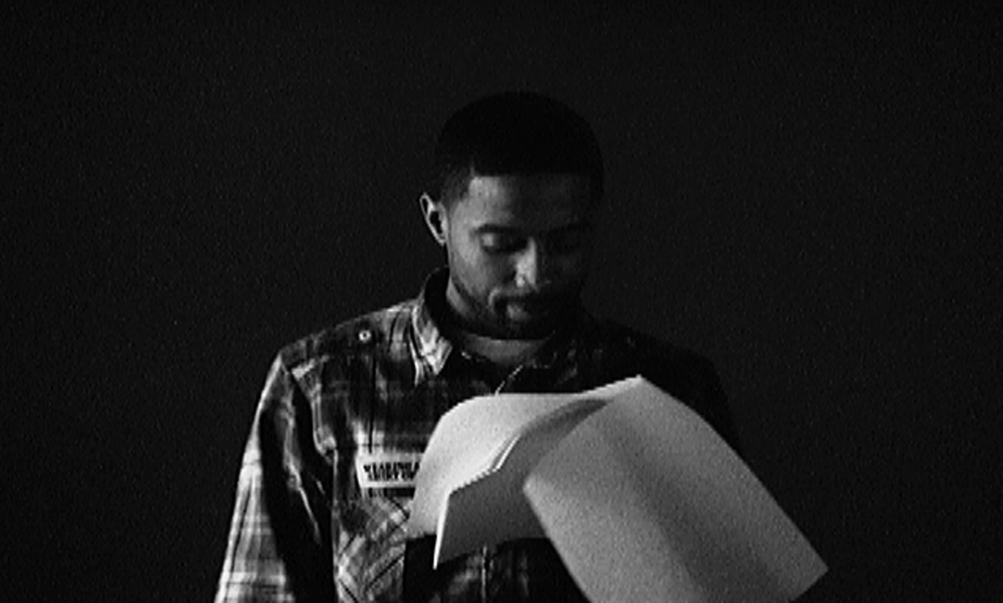A black and white photo of a man in a plaid shirt looking down at a sheet of paper he is holding. The background is dark, and the image has a grainy texture, giving it a vintage feel.