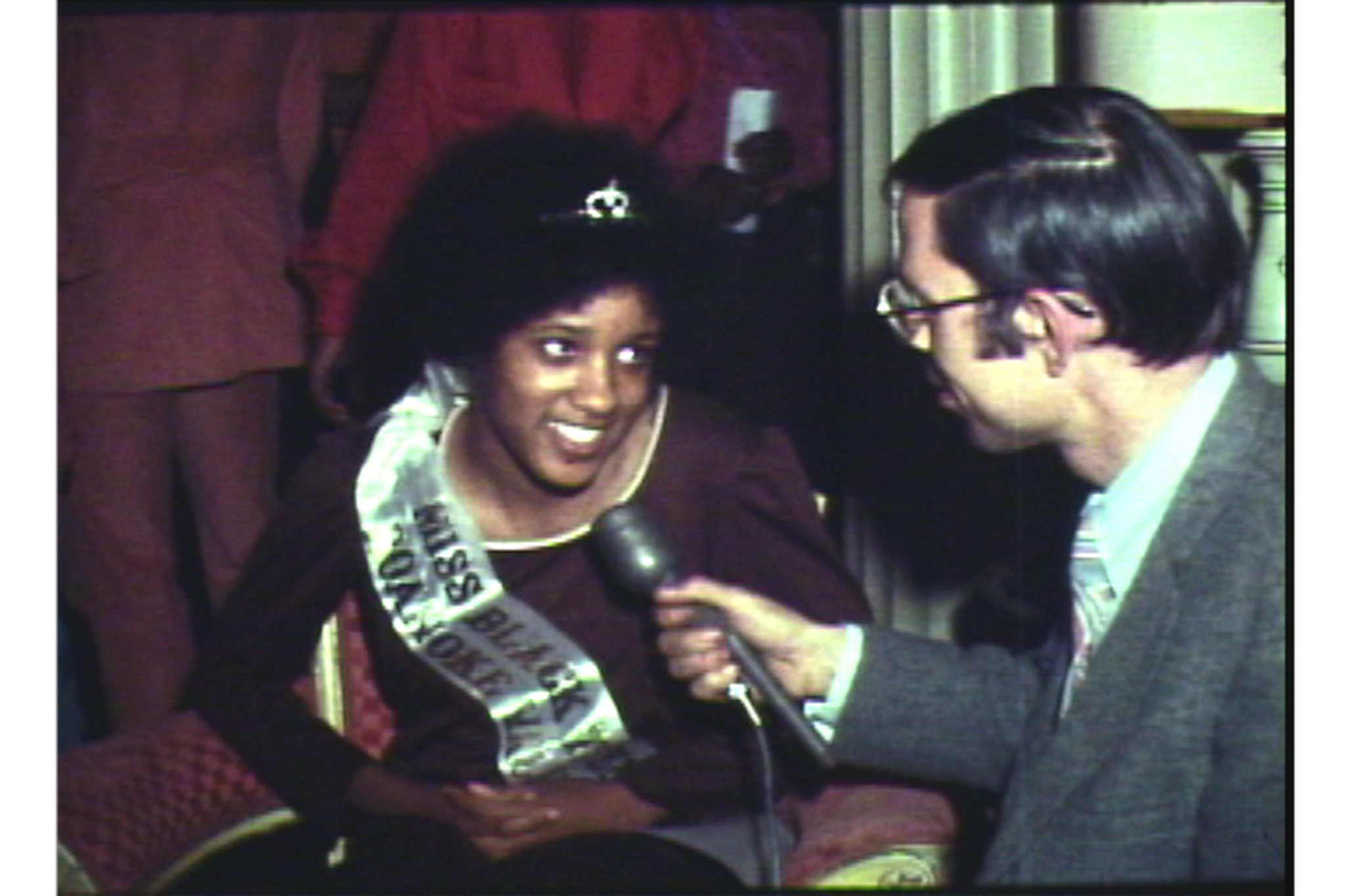 A woman wearing a sash that reads "Miss Black America" is being interviewed by a man holding a microphone. She has a bright smile and is looking slightly away from the interviewer, who is dressed in a suit and glasses. The setting appears to be indoors with a red backdrop.