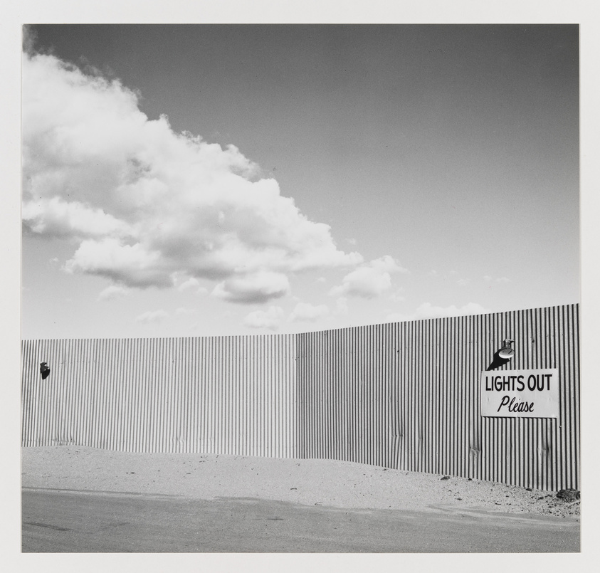 A corrugated metal fence with a sign that says "LIGHTS OUT Please" under a partly cloudy sky.