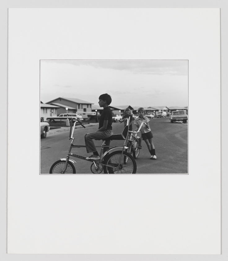 A black and white photograph capturing a moment from the past where a young boy sits atop a tall, custom bicycle, looking into the distance. Behind him, a girl is walking with a bicycle, both are in a residential area with houses and parked cars in the background. The image is mounted on a white border, giving it a framed appearance.