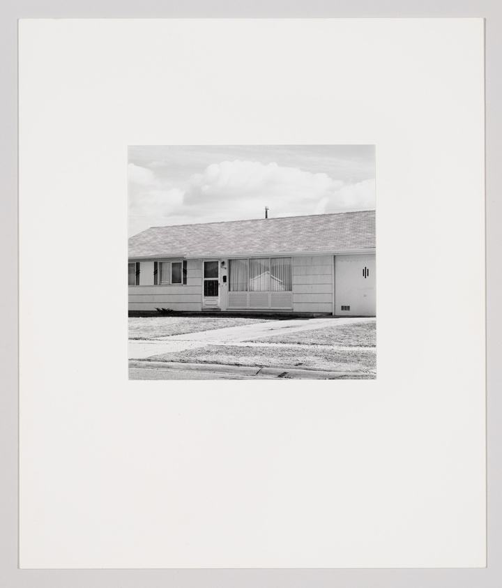 Single-story house with a front lawn, large window, and attached garage under a cloudy sky.