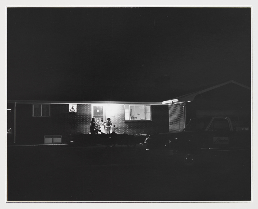 A black and white nighttime photo of a single-story house with a lit window where two people appear to be sitting at a table. In the foreground, there's a parked car with a visible nameplate, partially illuminated by the house's light. The scene is quiet and still, with a stark contrast between the darkness of the night and the brightness of the window.