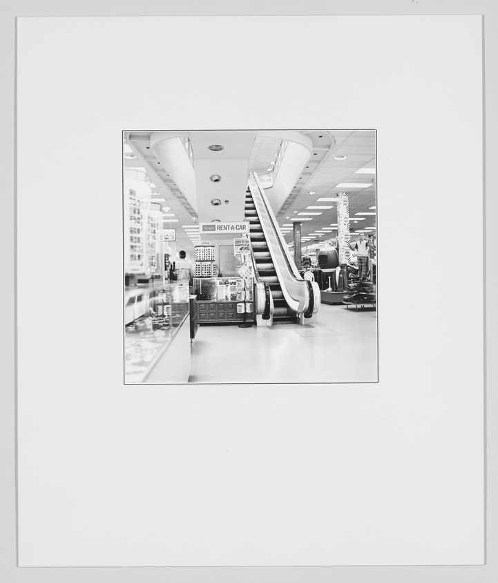 A black and white photograph of an escalator inside a modern store with various products on shelves, a "Rent-A-Car" sign visible, and a bright, well-lit ceiling with recessed lighting. The photo is framed within a larger white border.