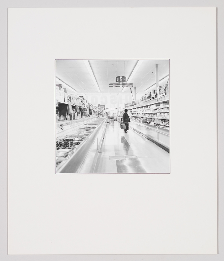 A black and white photograph of a person standing in the middle of a supermarket aisle, with shelves stocked with goods on either side and a sign indicating aisle number 5A overhead. The image is framed with a wide white border.