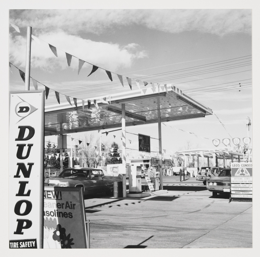 Gas station with cars parked at pumps, Dunlop tire safety sign, and promotional banners overhead.