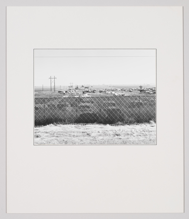 A chain-link fence in front of a field with many parked cars and distant power lines.