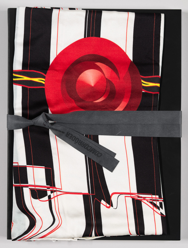 A folded graphic t-shirt with a bold red circular design on a black and white striped background, displayed on a gray surface. A black tag with white lettering is attached to the shirt.