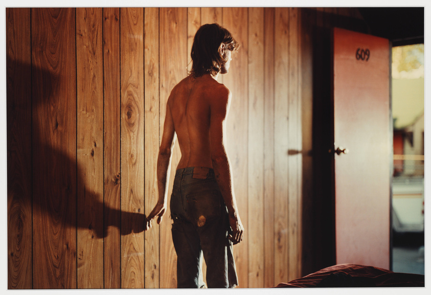 A shirtless man with long hair stands facing a wooden wall and a partially open door with the number 609 on it, sunlight casting his shadow on the wall. He is wearing worn jeans with a hole on the back and appears to be in a contemplative or waiting stance.