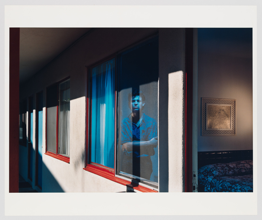 Person with arms crossed stands behind a window, reflected in the glass, next to a dimly lit room.