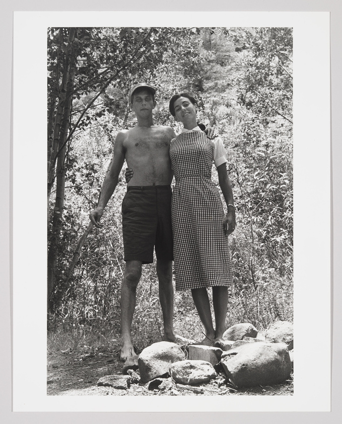 A black and white photograph of a shirtless man and a woman in a gingham dress standing close together on a rocky ground with trees and foliage in the background. The man has his left arm around the woman, and they both have a relaxed posture with a serene expression on their faces.