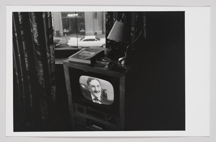 A black and white photo depicting a vintage television set in a dark room, displaying a smiling man in a suit. On top of the TV, there's a desk lamp, a book, and a typewriter. Heavy patterned curtains partially obscure the view of a window that looks out onto a street scene with a parked car.