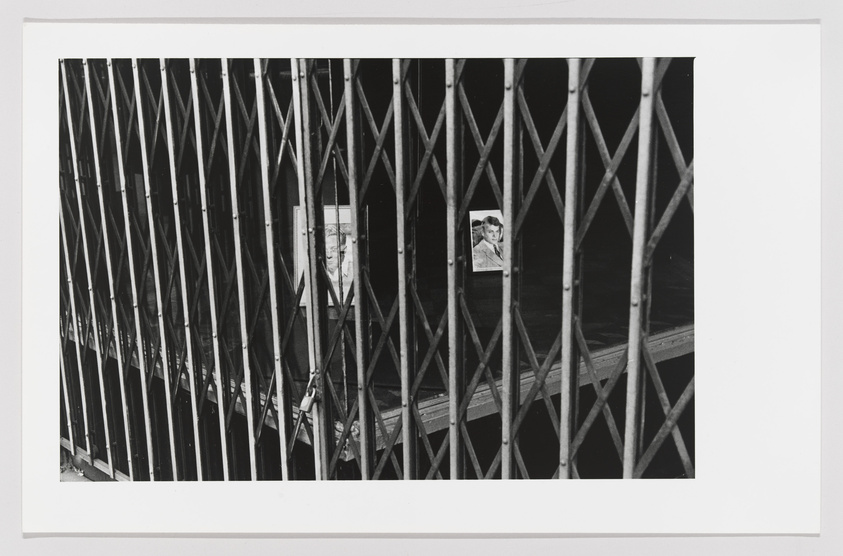 A black and white photograph showing a close-up view of a retractable metal gate with diamond-shaped patterns. Through the gaps of the gate, two framed portraits of individuals are partially visible, hanging on a wall in the background.