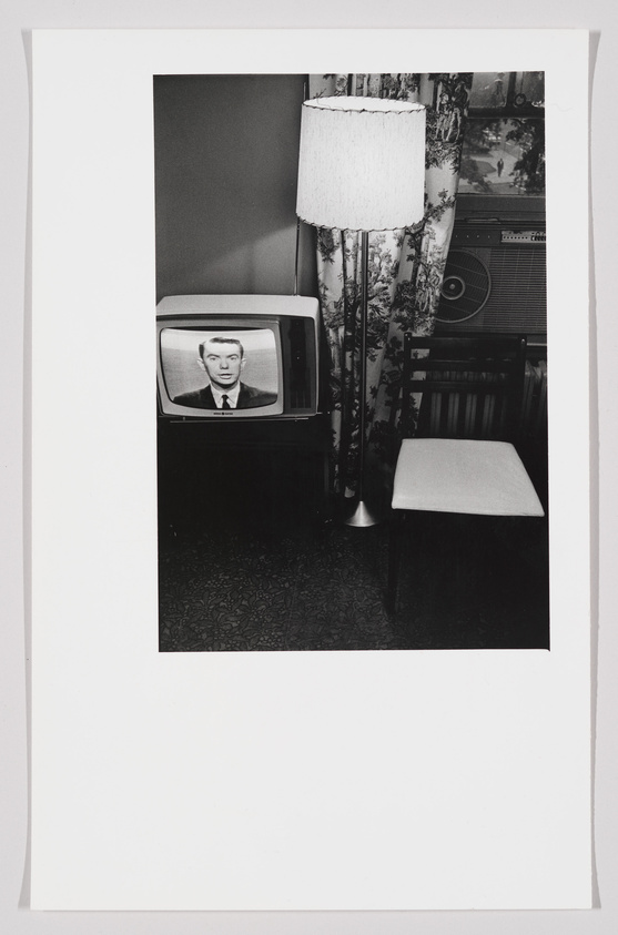 A black and white photograph depicting a vintage television set displaying a man's face, next to a floor lamp with a fringed shade, and a patterned curtain in the background. A wooden chair and a part of an air conditioning unit are also visible. The scene suggests a mid-20th-century living room setting.