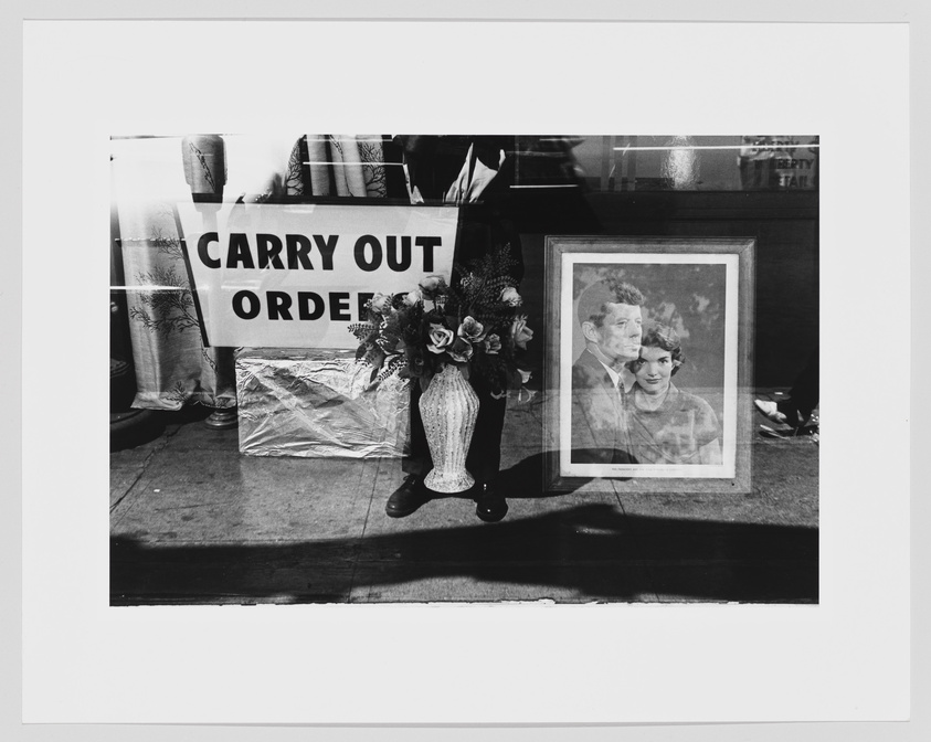 A black and white photograph featuring a framed portrait of a man and a woman, with a sign reading "CARRY OUT ORDERS" partially obscured behind it. In the foreground, a vase with artificial flowers is visible, and the reflection of a person can be seen on the glass covering the portrait. The setting appears to be a sidewalk or outdoor market.