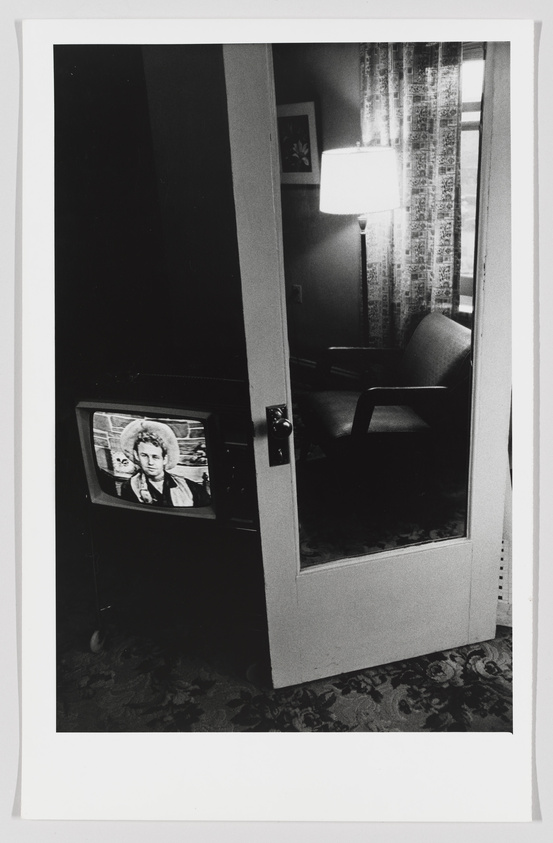 A black and white photograph showing a partially open door leading into a room with a vintage television set on, displaying a person's face. To the right, there's a lamp on a table illuminating patterned curtains beside a window, with a chair visible in the softly lit interior. The carpeted floor extends from the foreground into the room, featuring a floral design.